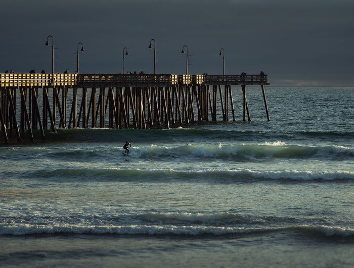 A surfer riding a wave beside Pismo Pier in early morning light, Pismo Beach, California